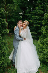 Portrait of the bride and groom standing on the background of green trees, embracing. The bride smiles sincerely. Stylish groom. Fashion and style. Beautiful bride