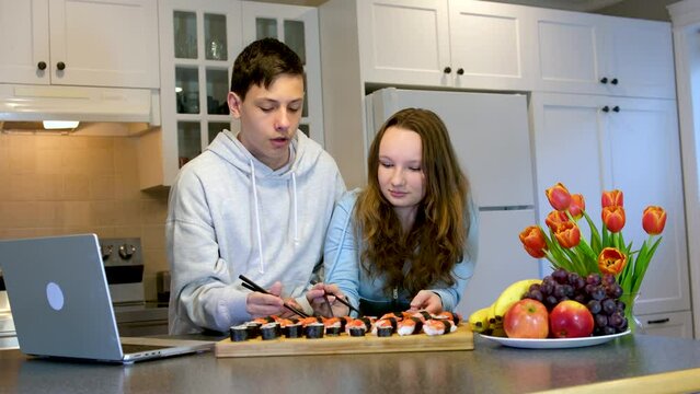 Attractive happy young couple eating sushi at home Romantic date of couple eating seafood teen boy and girl eating sushi in the kitchen looking at laptops film Flowers and fruits on the table