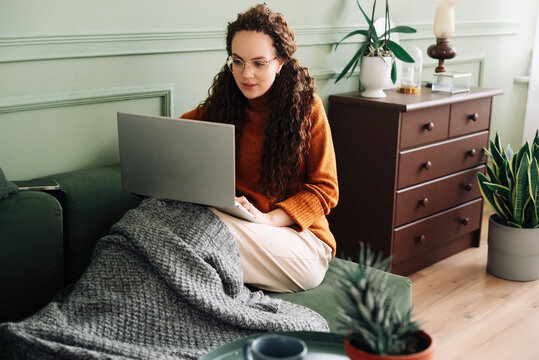 Relaxed young woman sitting on sofa holding mobile phone using cellphone and laptop technology doing online ecommerce shopping, texting messages relaxing on couch in cozy living room home interior.