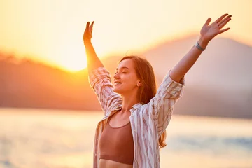 Tuinposter Portrait of calm happy smiling free woman with open arms and closed eyes enjoys a beautiful moment life on the seashore at sunset time © Goffkein
