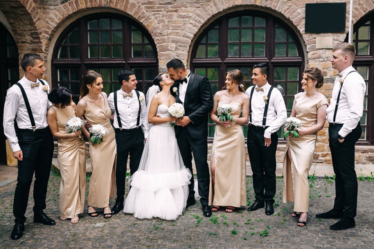 Handsome brunette groom kissing beautiful bride in wedding dress with bridesmaids bouquet and friends in the background