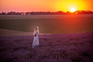 Fototapeta na wymiar Young blonde woman stands in purple field at sunset in the bright rays of sun. Travel in countryside. Allergy concept. Sunset sky. Carefree girl with long healthy hair. human countryside lifestyle