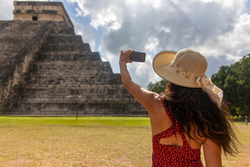 Fototapeta premium Sexy tourist girl with hat in front of the background of the pyramid of Kukulcan in the Mexican city of Chichen Itza taking a photo with mobile phone. Travel concept.