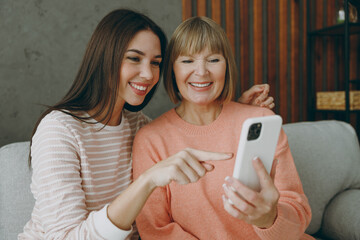 Two adult smiling women mature mom young kid wearing casual clothes hold use mobile cell phone sit on gray sofa couch stay at home flat rest relax spend free spare time in living room. Family concept.