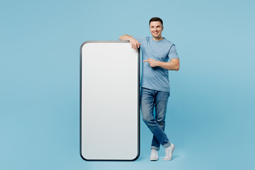 Full body young happy man wear casual t-shirt point index finger on big huge blank screen mobile cell phone with workspace copy space mockup area isolated on plain pastel light blue cyan background.