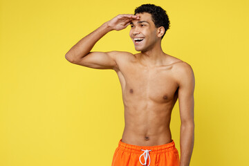 Fototapeta premium Young sexy smiling fun man wear orange shorts swimsuit hold hand at forehead look far away distance relax near hotel pool isolated on plain yellow background. Summer vacation sea rest sun tan concept.