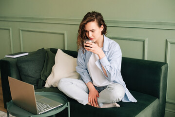 Adorable female drinking hot tea sitting on a sofa near a big window and watching webinars and...
