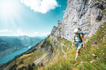 Fototapeta premium Atheltic woman hiking on flowery trail next to steep rock wall with scenic view on Walensee and Churfürsten mountain range in the background. Schnürliweg, Walensee, St. Gallen, Switzerland, Europe.