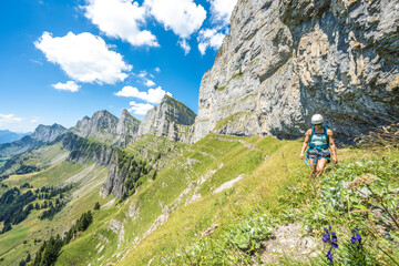 Fototapeta na wymiar Mountaineer hiking on flowery meadow below steep rock wall with scenic view on the Churfürsten mountain range. Schnürliweg, Walensee, St. Gallen, Switzerland, Europe.