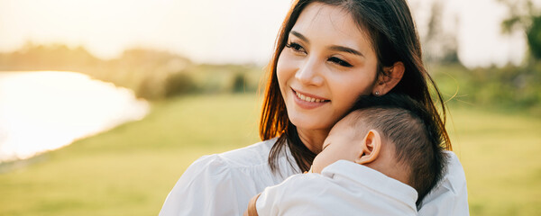 Obraz premium Family care. Happy beautiful mother holding baby sleeping son in the park at evening time before sunset, Portrait of mom hold newborn feeling happy and smiles in the garden nature outdoor, Mother Day