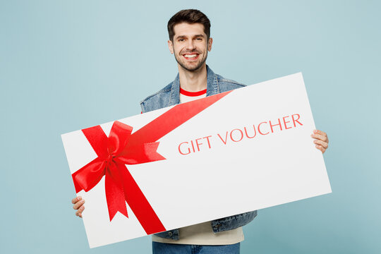 Young fun man he wear denim vest red t-shirt casual clothes hold huge large gift certificate coupon voucher card for store isolated on plain pastel light blue cyan background studio. Lifestyle concept