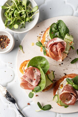 Bruschetta with ham or prosciutto, cream cheese, cucumber, spinach and microgreens. Tasty sandwich or toasted bread on white tray. Appetizer or breakfast, top view, flat lay