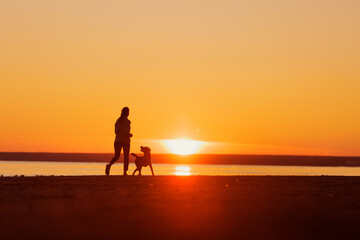 woman and dog on run on seashore, silhouette against background of setting sun. healthy lifestyle, freedom and outdoor sports. labrador retriever dog
