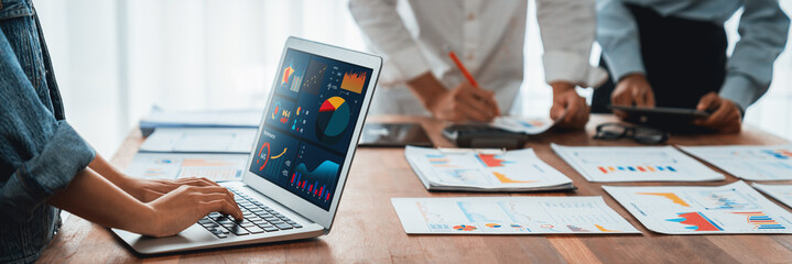 Panorama shot analyst team utilizing BI Fintech to analyze financial report with laptop. Businesspeople analyzing BI power dashboard displayed on laptop screen for business insight. Scrutinize