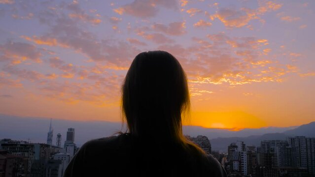 Woman is standing on the balcony and looking around against the sunset, sunrise sky over the city - central composition, back view. Lonely, urban, dramatic and freedom concept