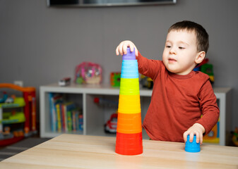 A cute little toddler boy of two years old sits at a children's table and plays with multi-colored pyramids in his room. Educational toys for shildren. Soft selective focus