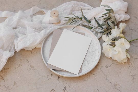 Summer wedding stationery. Birthday mock-up scene. Blank greeting cards on plate. White oleander blossom, ribbon and olive tree branches. Beige marble background, floor. Flat lay, top view.