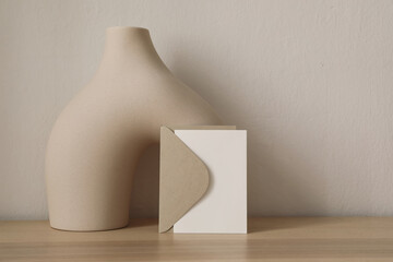 Closeup of blank greeting card, invitation mockup with craft envelope. Beige boho ceramic vase,...