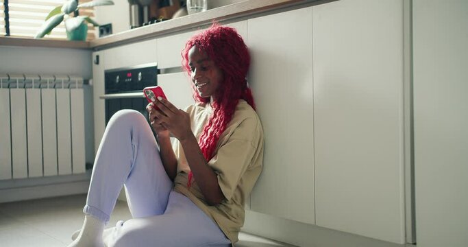 Joyful African American girl with red curly hair sitting on kitchen floor and srfing internet using mobile