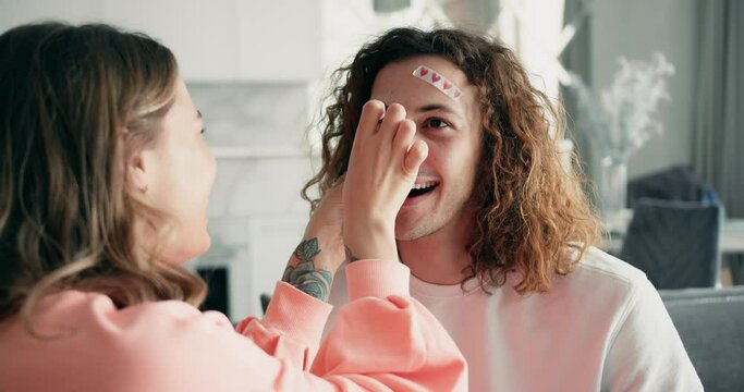 Close up of cheerful excited careless happy with toothy beaming smile man with curly hair and woman stick funny stickers on his face