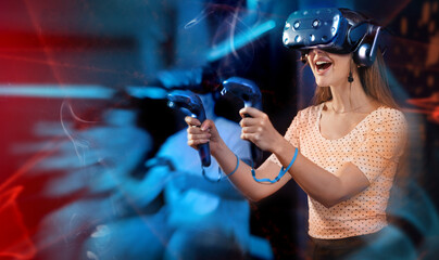 Cheerful emotional young woman in VR headset fully immersed in game, actively engaged in virtual reality world, manipulating objects or shooting targets with joysticks in hands.. © JackF