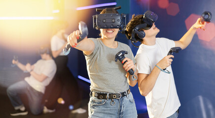 Excited modern young couple with gaming controllers in hands and VR goggles having fun with friends in virtual reality room. Concept of immersive interactive experience..