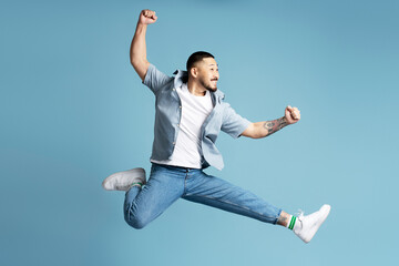 Happy emotional asian man wearing stylish casual clothes, white shoes running fast, jumping high isolated on blue background. Positive lifestyle concept 