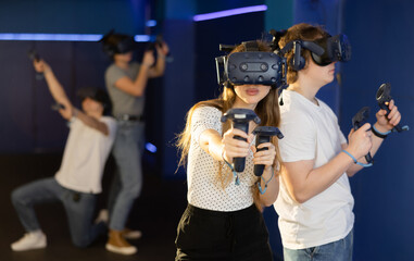 Two teenagers in virtual reality glasses with joysticks stand shoulder to shoulder and play...