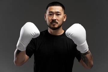 Portrait of serious handsome asian boxer wearing white boxing gloves looking at camera, isolated on black background. Sport competition, motivation concept 