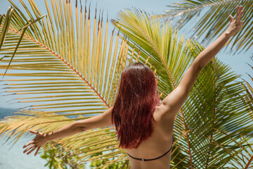 Young beautiful tanned girl in bikini stands with her back with hands up against the background of palm trees on the beach of exotic island against background of azure ocean and sky. Travel, vacation