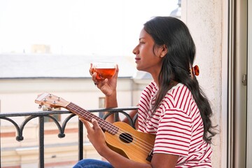 Indian woman playing ukulele on windowsill drinking a tea cup