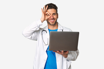 Young doctor man holding a laptop excited keeping ok gesture on eye.