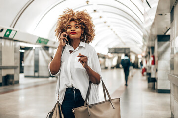 Beautiful fashionable black woman standing at a subway train station. She is happy and talking to someone on her smart phone. Public transportation and urban life concept. - obrazy, fototapety, plakaty