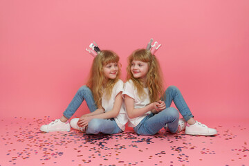 Obraz na płótnie Canvas Twin girls looking at each other. Two beautiful and positive curly blonde sisters who are 6 or 7 years old, wearing white T-shirts, jeans, and festive crowns sitting on a pink background with confetti