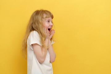 The portrait of a happy, excited, and astonished child. A shocked curly-haired girl in a white T-shirt with an open mouth on an isolated yellow background with space for text. 