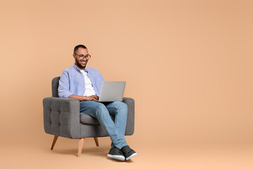 Smiling young man working with laptop in armchair on beige background, space for text