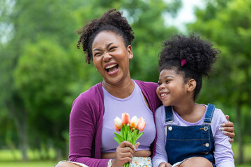 African mother holding bouquet of tulip flowers and the daughter hugging each other while happily enjoying picnic in the public park during summer for family love and care in mother's day celebration