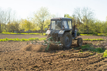 Fototapeta premium The tractor drives across the field and cultivates the land. Agricultural vehicle works in countryside. Sowing is the process of planting seeds in the ground as part of the early spring time