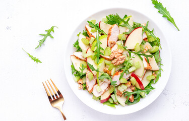 Waldorf salad with red and green apple, celery, lettuce, chicken fillet, arugula and walnuts on plate, white table background, top view