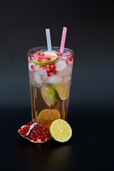 A tall glass of refreshing lemonade with pomegranate seeds and ice on a black background, next to ripe fruits.