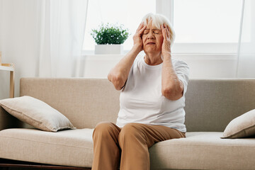 Elderly woman severe pain in the head sitting on the couch, health problems in old age, poor...
