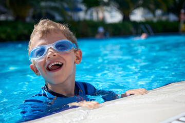 Satisfied boy swims in a warm summer pool in blue clear water wearing swimming glasses. A cheerful...