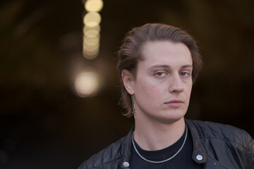 A genderfluid (LGBTQ) model poses outside, in front of a tunnel, to express calmness and a sense of overcoming challenges. Their hair is combed back, and they are wearing a black leather jacket.