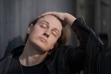 A genderfluid model (he, she, they) wears a black leather jacket and stands in front of a gray wall, running their hand through their hair.