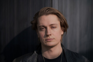 A genderfluid model (he, she, they) wears a black leather jacket and stands in front of a gray wall, looking straight at the camera to express pride and queer identity.