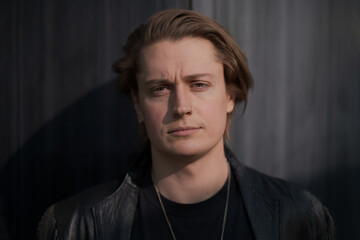 Fototapeta premium A genderfluid model (he, she, they) wears a black leather jacket and stands in front of a gray wall, looking straight at the camera to express pride and queer identity.
