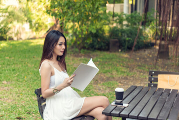 Asian woman holding book reading magazine at green park in natural garden. Young woman relaxation read open book self study. University women smiling with happiness learning. Women sit in green park