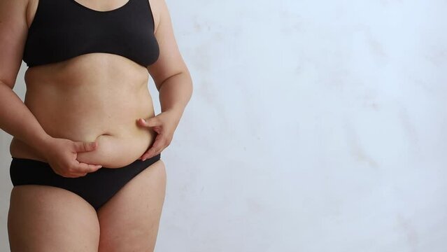 Unrecognizable overweight woman wearing black underwear, lifting excessive fat of stomach with hands, doing massage on white background. Weight loss, body care, plastic surgery. Copy space, banner.