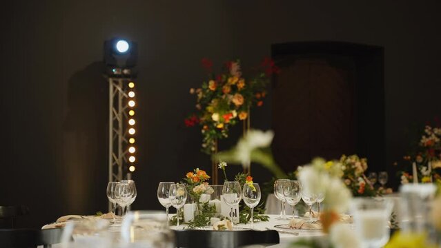 Served tables in dark walled banquet hall, decorated with floral arrangements and illuminated by professional lighting equipment before event. Spotlight turns its head and beam illuminates hall.