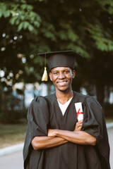 Smiling afro american graduate from university stand outdoors in black mantle and hat with crossed...